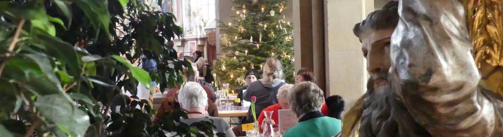 links eine Pflanze, rechts die Mose-Figur unter der Kanzel. Dazwischen blickt man auf essende Menschen, im Hintergrund der Weihnachtsbaum.