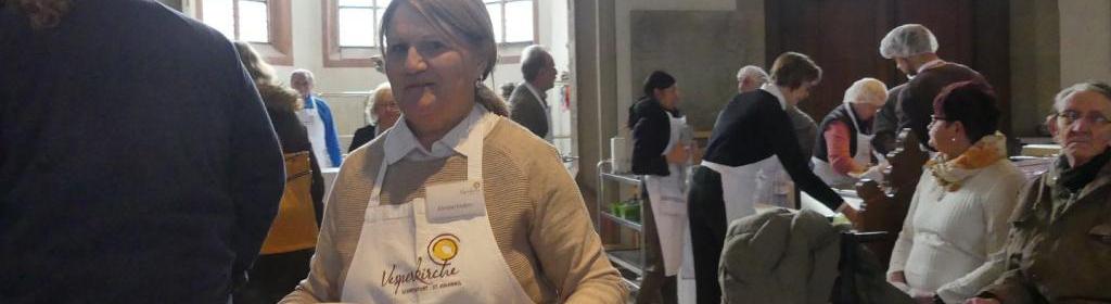 Christel Enders in Vesperkirche-Schürze. Sie trägt ein Tablett mit zwei Tellern mit Braten sowie zwei Salatschüsseln.